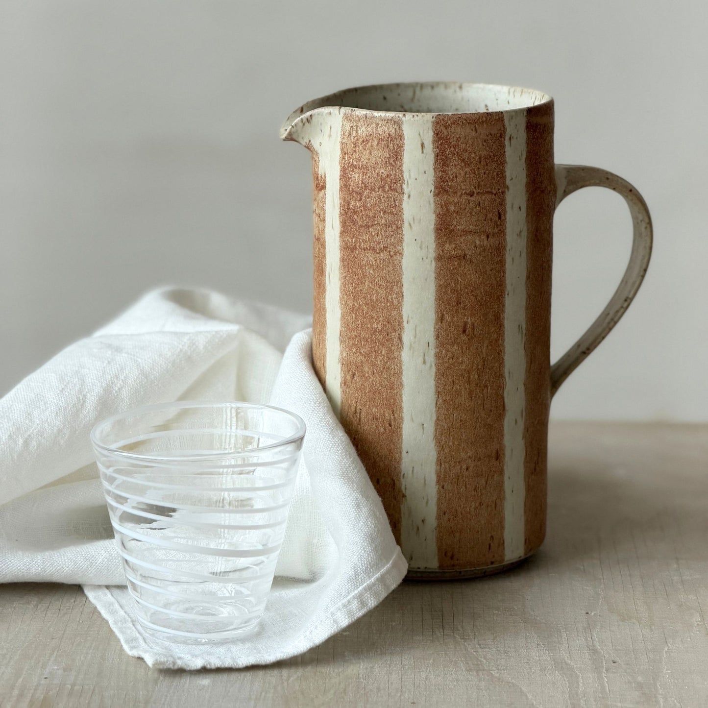 Handgjord randig keramikkanna i brun glasyr på havrefärgad botten med ett randigt glas från Paradisverkstaden, cirka 20 cm hög.